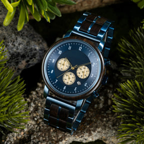A chronograph watch with a combination of wooden and blue metal bracelet, featuring a blue dial with beige chronograph sub-dials, numerical hour markers, and a date window, placed on a rock surrounded by green foliage.