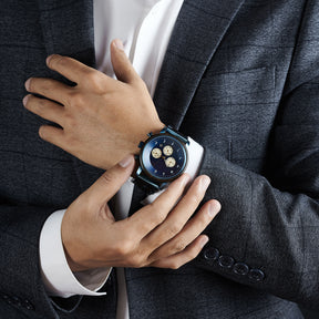 A man wearing a grey suit and white shirt, showcasing a chronograph watch with a combination of wooden and blue metal bracelet on his wrist, featuring a blue dial with beige chronograph sub-dials and numerical hour markers, against a dark background.