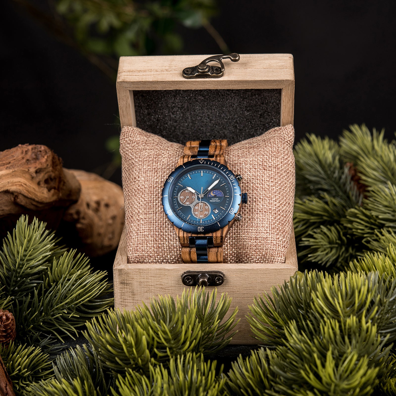 A multifunction watch with a combination of wooden and blue metal bracelet, featuring a blue dial with chronograph sub-dials, moon phase display, water-resistant label, and date window, placed inside a wooden box with a burlap pillow, surrounded by green foliage and natural driftwood elements.