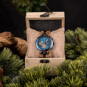 A multifunction watch with a combination of wooden and blue metal bracelet, featuring a blue dial with chronograph sub-dials, moon phase display, water-resistant label, and date window, placed inside a wooden box with a burlap pillow, surrounded by green foliage and natural driftwood elements.