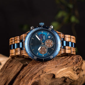 A multifunction watch with a combination of wooden and blue metal bracelet, featuring a blue dial with chronograph sub-dials, moon phase display, water-resistant label, and date window, placed on a piece of driftwood surrounded by green foliage.