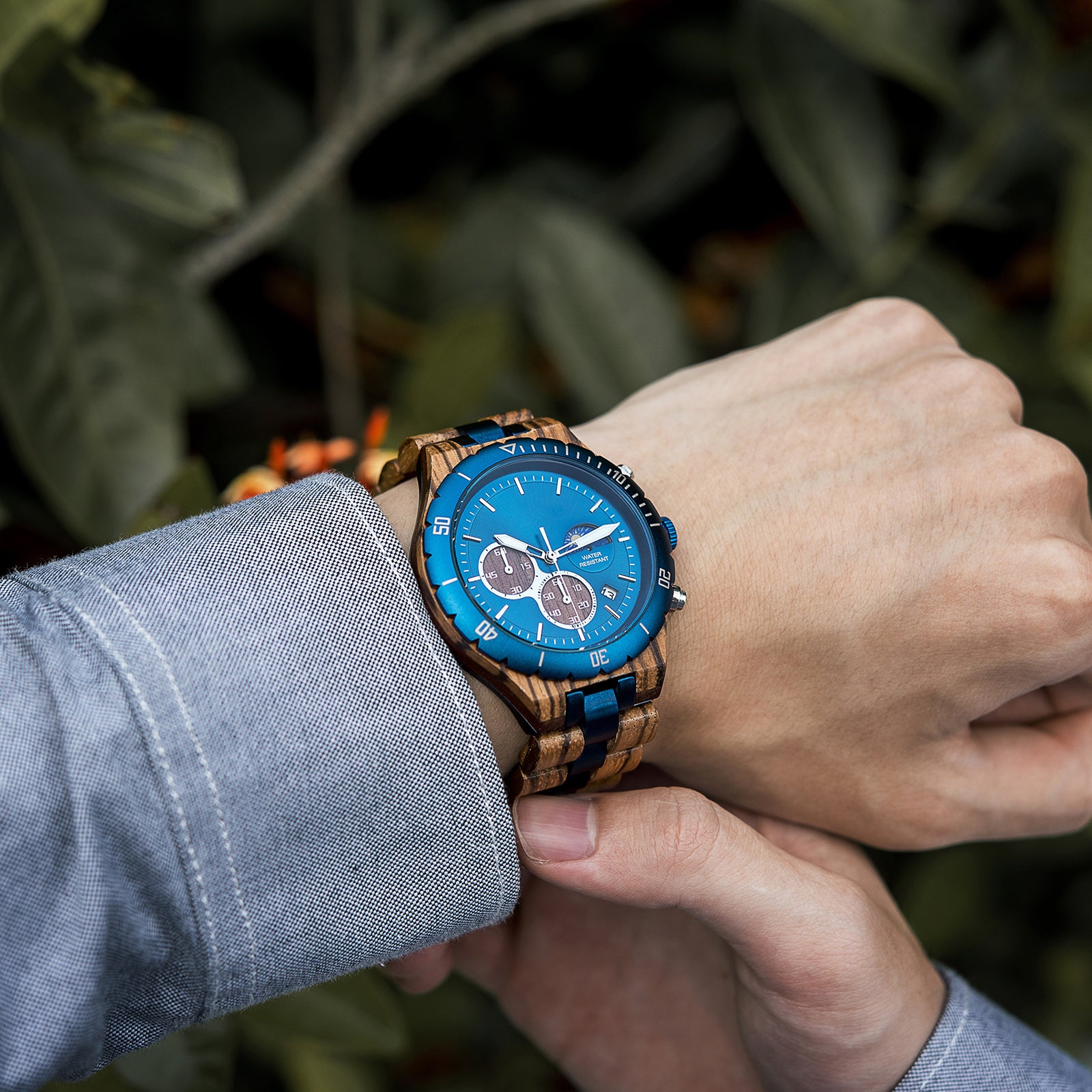 A man wearing a grey shirt, showcasing a multifunction watch with a wooden and blue metal bracelet on his wrist, featuring a blue dial with chronograph sub-dials, moon phase display, water-resistant label, and date window, against a background of green foliage and orange flowers.