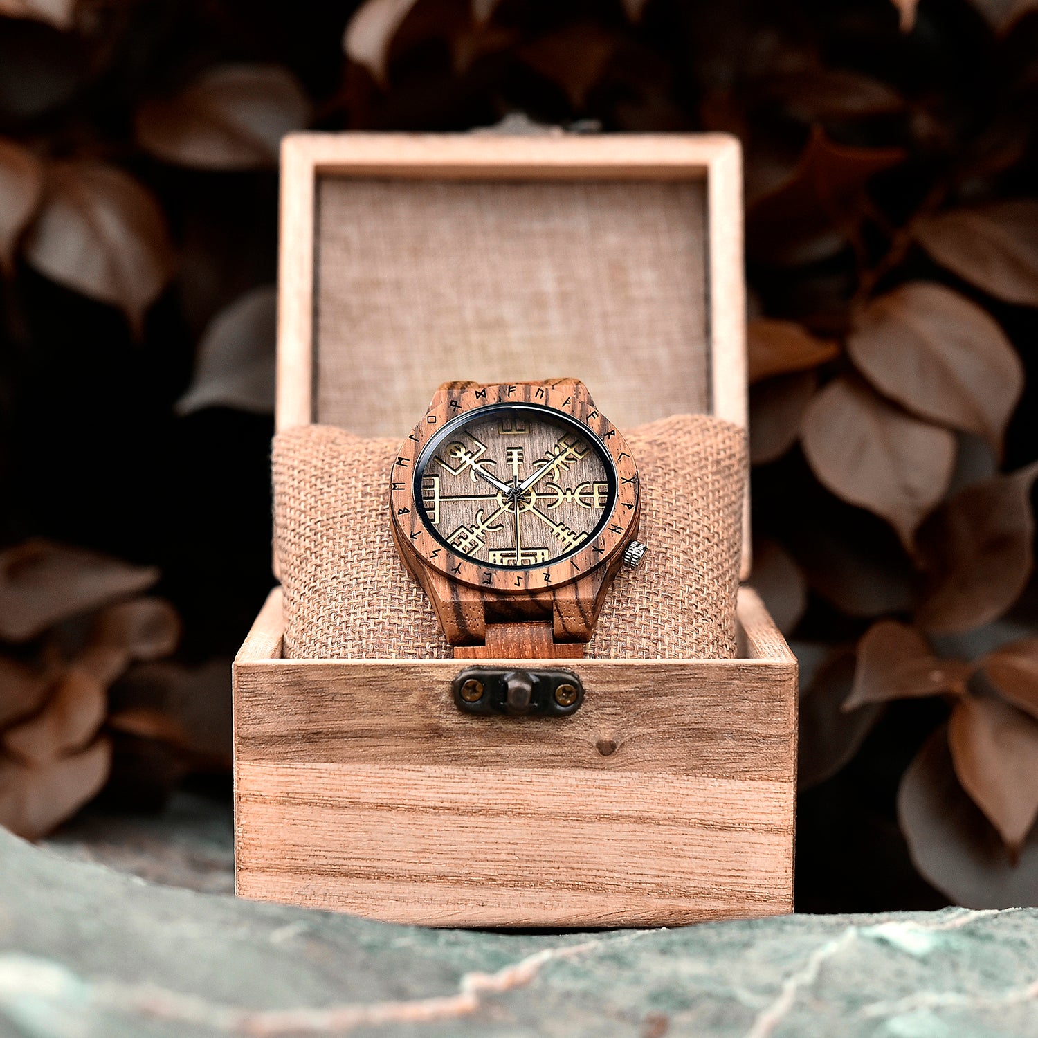 A brown wooden wristwatch with golden Viking symbols and runes, placed on a burlap cushion inside a wooden box, surrounded by brown foliage.
