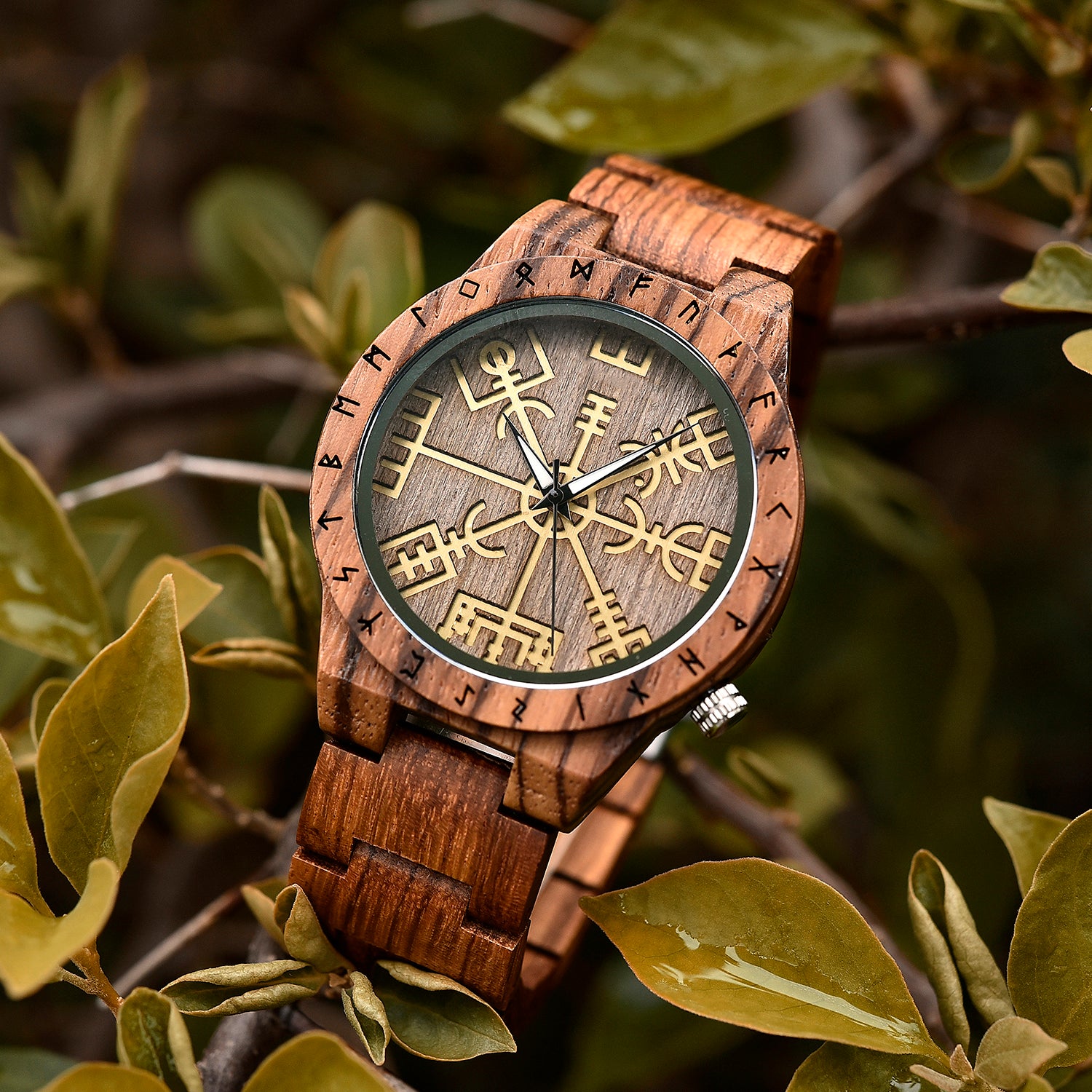 A brown wooden wristwatch with golden Viking symbols and runes on the dial, placed on green foliage with water droplets on the leaves.