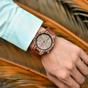 A person wearing a brown wooden wristwatch with golden Viking symbols and runes on the dial, paired with a light blue shirt, against a backdrop of palm leaves.