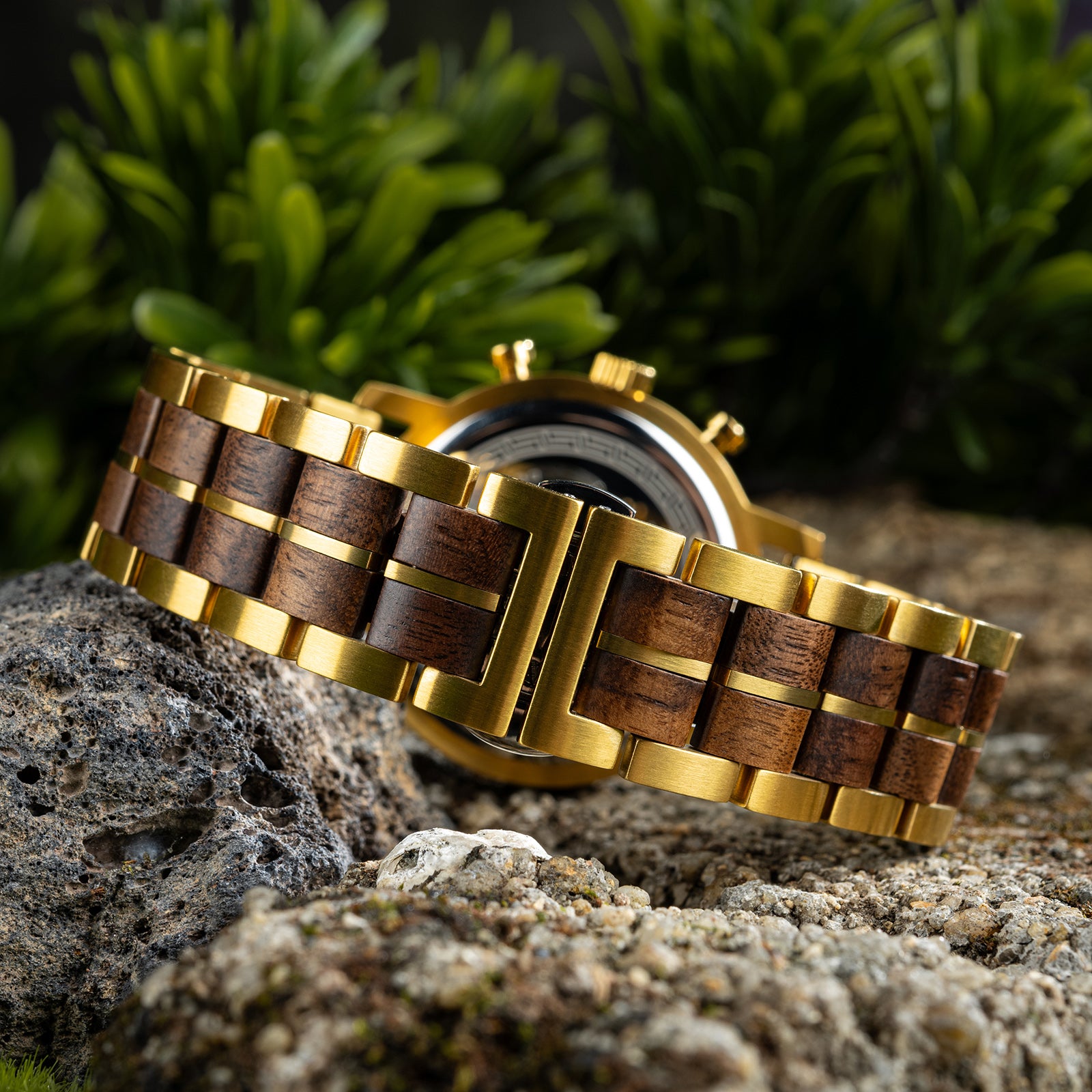 The case back of a chronograph watch with a combination of wooden and gold metal bracelet, featuring an engraved Greek key pattern, placed on a piece of volcanic rock with green foliage in the background.