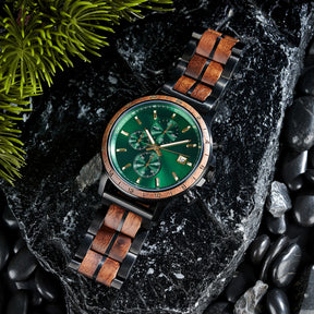 An angled close-up of a chronograph watch with a combination of black metal and light wood bracelet, featuring a green dial with multiple sub-dials, 24-hour markers on the bezel, and a date window, placed on black rock with green foliage in the background.