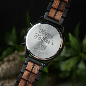 A close-up of the case back of a chronograph watch with a combination of black metal and light wood bracelet, showcasing its polished finish, set against green foliage in the background.

