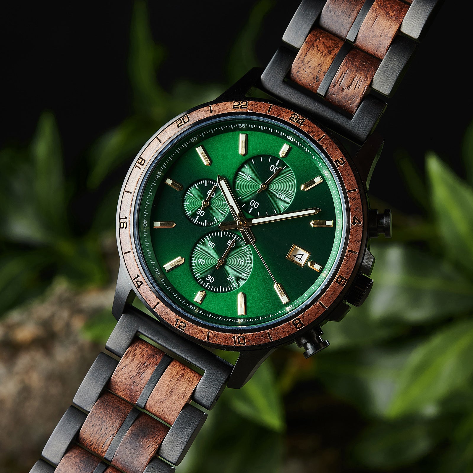 A close-up of a chronograph watch with a combination of black metal and light wood bracelet, featuring a green dial with multiple sub-dials, 24-hour markers on the bezel, and a date window, set against green foliage in the background.