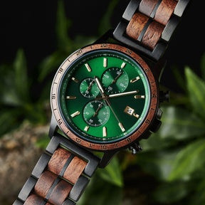 A close-up of a chronograph watch with a combination of black metal and light wood bracelet, featuring a green dial with multiple sub-dials, 24-hour markers on the bezel, and a date window, set against green foliage in the background.