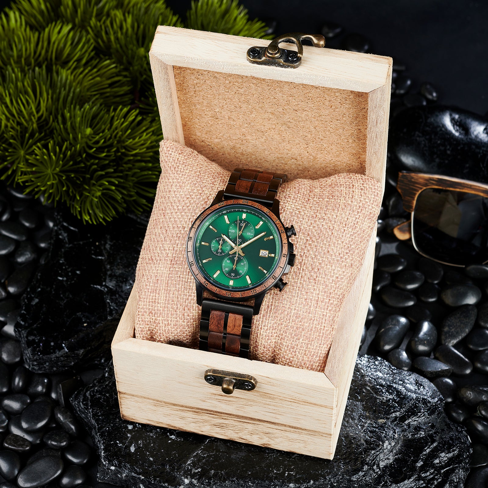 A chronograph watch with a combination of black metal and light wood bracelet, featuring a green dial with multiple sub-dials, 24-hour markers on the bezel, and a date window, placed inside a wooden box with a burlap pillow, surrounded by black rocks and green foliage.
