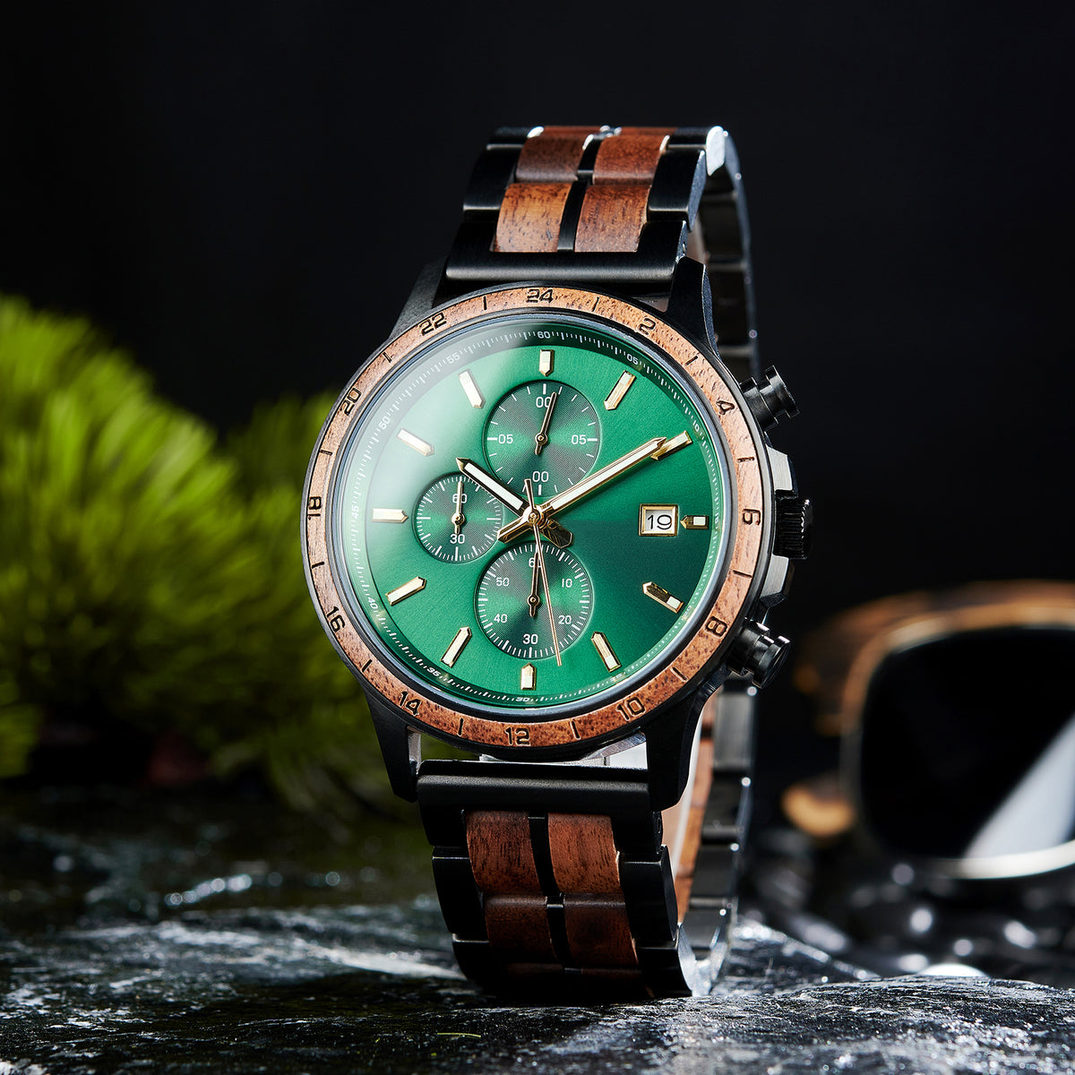 A chronograph watch with a combination of black metal and light wood bracelet, featuring a green dial with multiple sub-dials, 24-hour markers on the bezel, and a date window, placed on wet rock with green foliage in the background.