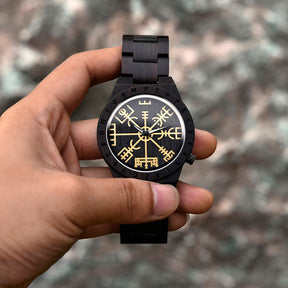 A hand holding a dark wooden wristwatch with golden Viking symbols and runes on the dial, set against a blurred natural outdoor background.