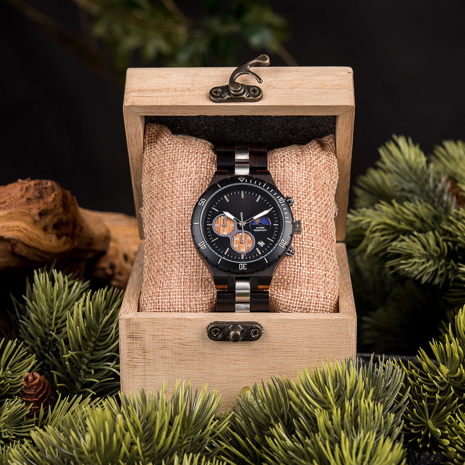 A multifunction watch with a wooden and metal bracelet, featuring a black dial with chronograph sub-dials, moon phase display, and water-resistant label, placed inside a wooden box with a burlap pillow, surrounded by green foliage and natural elements.