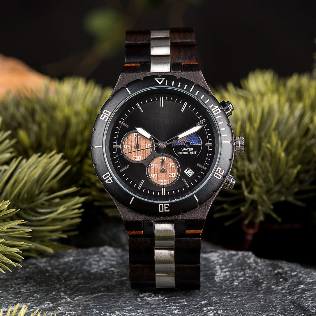 A multifunction watch with a combination of wooden and metal bracelet, featuring a black dial with chronograph sub-dials and water-resistant label, placed on a rock surrounded by green foliage.