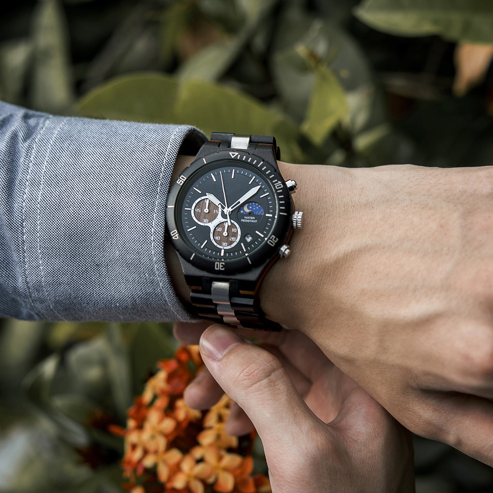 A man wearing a blue shirt, showcasing a multifunction watch with a wooden and metal bracelet on his wrist, featuring a black dial with chronograph sub-dials, moon phase display, and water-resistant label, against a background of green foliage and orange flowers.