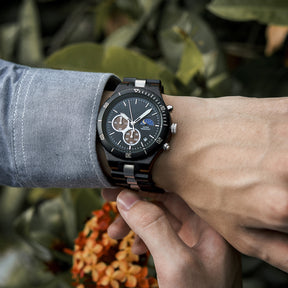 A man wearing a blue shirt, showcasing a multifunction watch with a wooden and metal bracelet on his wrist, featuring a black dial with chronograph sub-dials, moon phase display, and water-resistant label, against a background of green foliage and orange flowers.