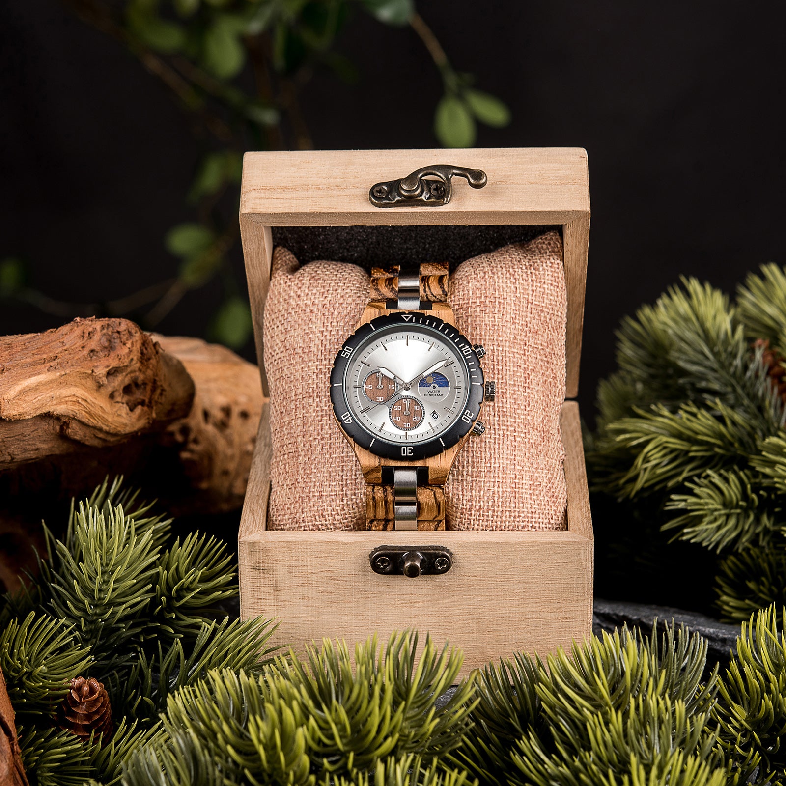 A multifunction watch with a combination of wooden and silver metal bracelet, featuring a silver dial with brown chronograph sub-dials, moon phase display, water-resistant label, and date window, placed inside a wooden box with a burlap pillow, surrounded by green foliage and natural driftwood elements.