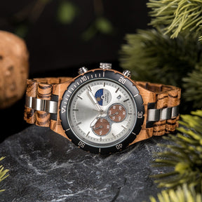 A multifunction watch with a combination of wooden and silver metal bracelet, featuring a silver dial with brown chronograph sub-dials, moon phase display, water-resistant label, and date window, placed on a black rock surrounded by green foliage and natural driftwood elements.