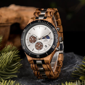 A multifunction watch with a combination of wooden and silver metal bracelet, featuring a silver dial with chronograph sub-dials, moon phase display, water-resistant label, and date window, placed on a rock surrounded by green foliage and natural driftwood elements.