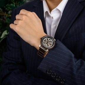 A man wearing a navy pinstripe suit and white dress shirt, showcasing a skeleton mechanical watch with a wooden and metal bracelet on his wrist.
