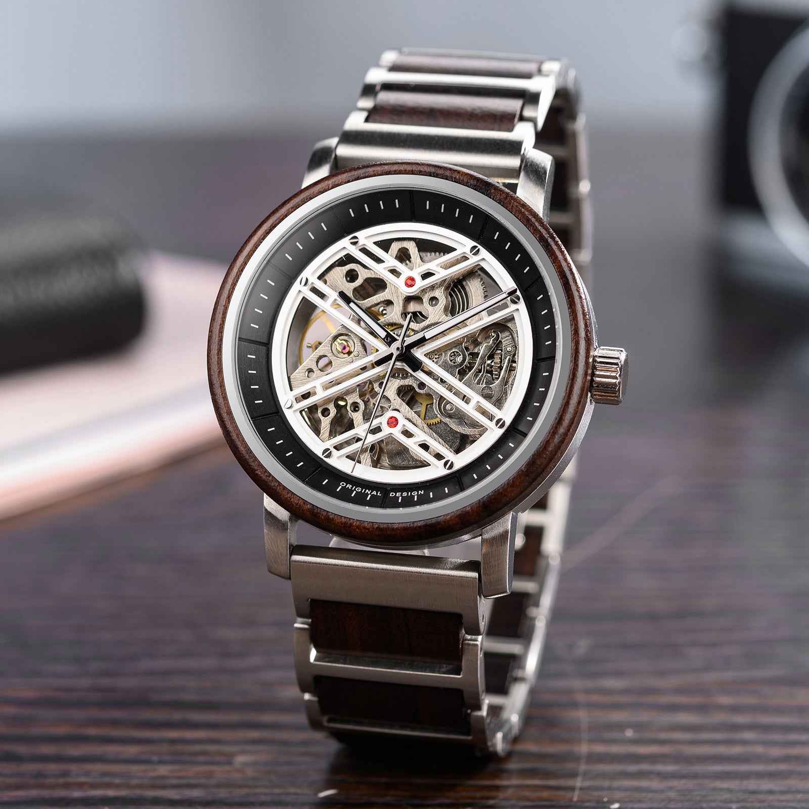 A detailed view of a skeleton mechanical watch with a combination of wooden and metal bracelet, featuring an open-work dial that reveals intricate movement, a dark wooden bezel, and stainless steel accents, placed on a wooden surface with blurred background elements.
