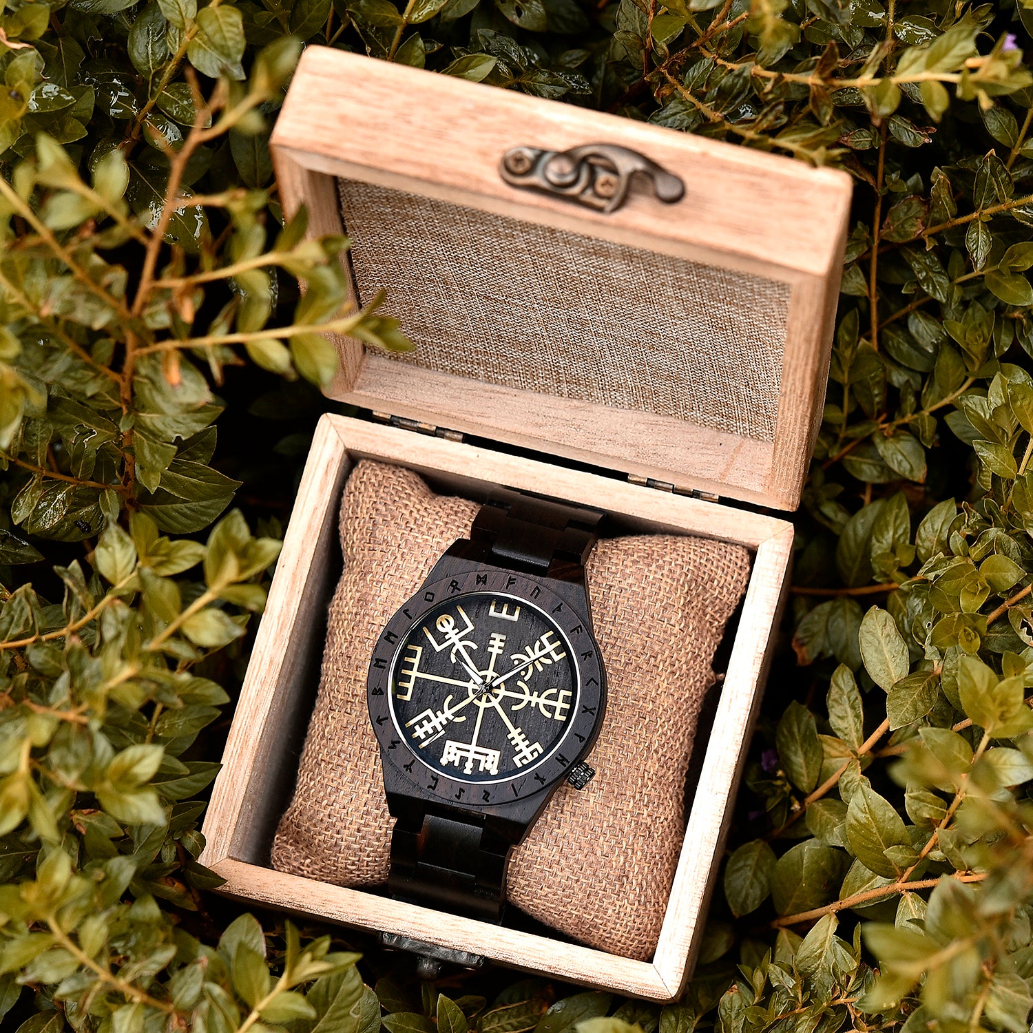 A dark wooden wristwatch with golden Viking symbols and runes, placed on a burlap cushion inside a wooden box, surrounded by green foliage.
