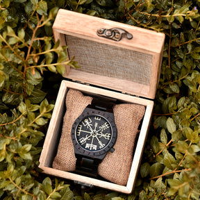 A dark wooden wristwatch with golden Viking symbols and runes, placed on a burlap cushion inside a wooden box, surrounded by green foliage.