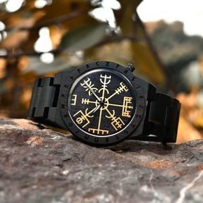 A black wooden wristwatch featuring golden Viking symbols and runes on the dial, placed on a rock with a blurred natural background of trees and foliage.
