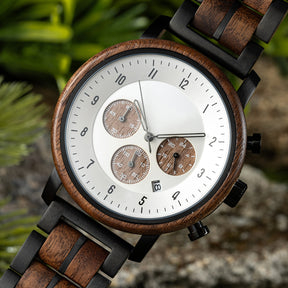 A close-up of a chronograph watch with a combination of wooden and black metal bracelet, featuring a white dial with brown chronograph sub-dials, numerical hour markers, and a date window, placed on a rock with green foliage in the background.