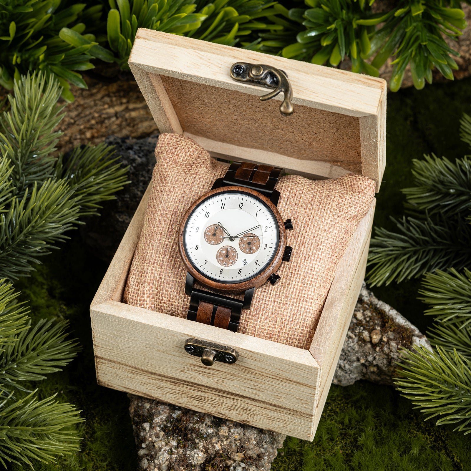 A chronograph watch with a combination of wooden and black metal bracelet, featuring a white dial with brown chronograph sub-dials and numerical hour markers, placed inside a wooden box with a burlap pillow, surrounded by green foliage and natural rock elements.