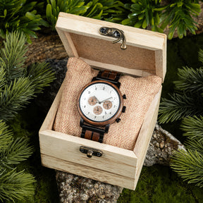 A chronograph watch with a combination of wooden and black metal bracelet, featuring a white dial with brown chronograph sub-dials and numerical hour markers, placed inside a wooden box with a burlap pillow, surrounded by green foliage and natural rock elements.