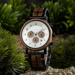 A chronograph watch with a combination of wooden and black metal bracelet, featuring a white dial with brown chronograph sub-dials, numerical hour markers, and a date window, placed on a rock surrounded by green foliage.