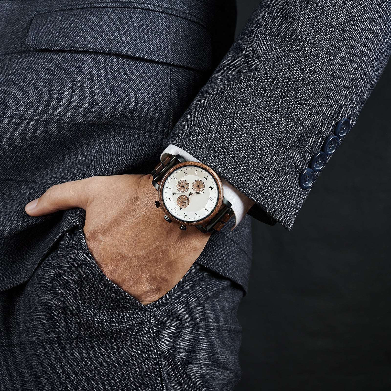 A man wearing a grey suit, showcasing a chronograph watch with a combination of wooden and black metal bracelet on his wrist, featuring a white dial with brown chronograph sub-dials and numerical hour markers, against a dark background.