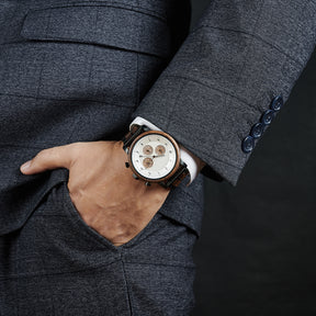 A man wearing a grey suit, showcasing a chronograph watch with a combination of wooden and black metal bracelet on his wrist, featuring a white dial with brown chronograph sub-dials and numerical hour markers, against a dark background.