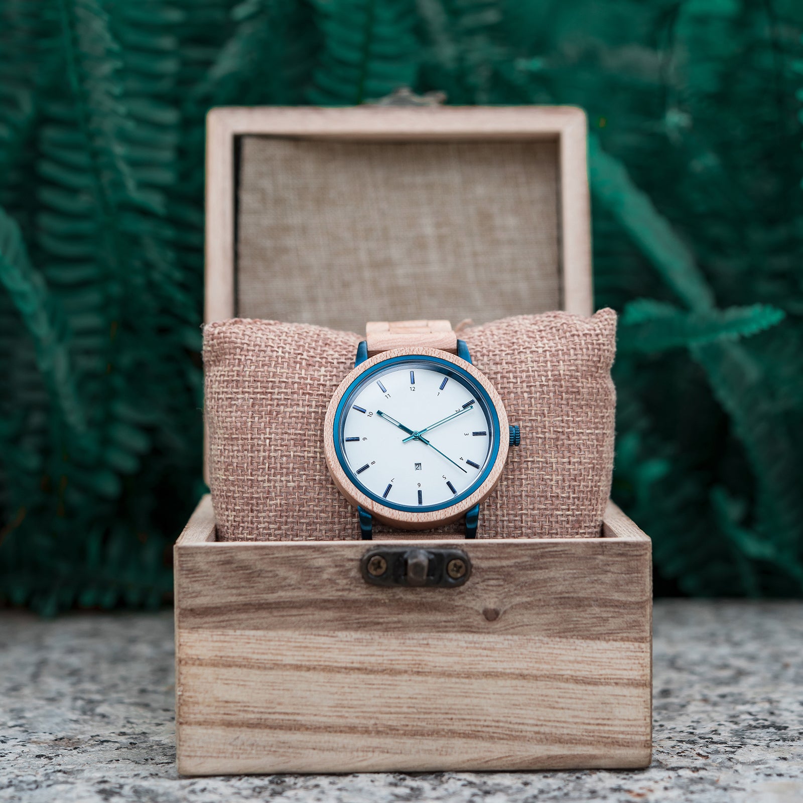 A wooden wristwatch with a white dial and blue accents, placed on a burlap cushion inside a wooden box, with green ferns in the background.
