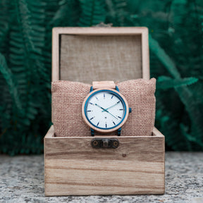 A wooden wristwatch with a white dial and blue accents, placed on a burlap cushion inside a wooden box, with green ferns in the background.

