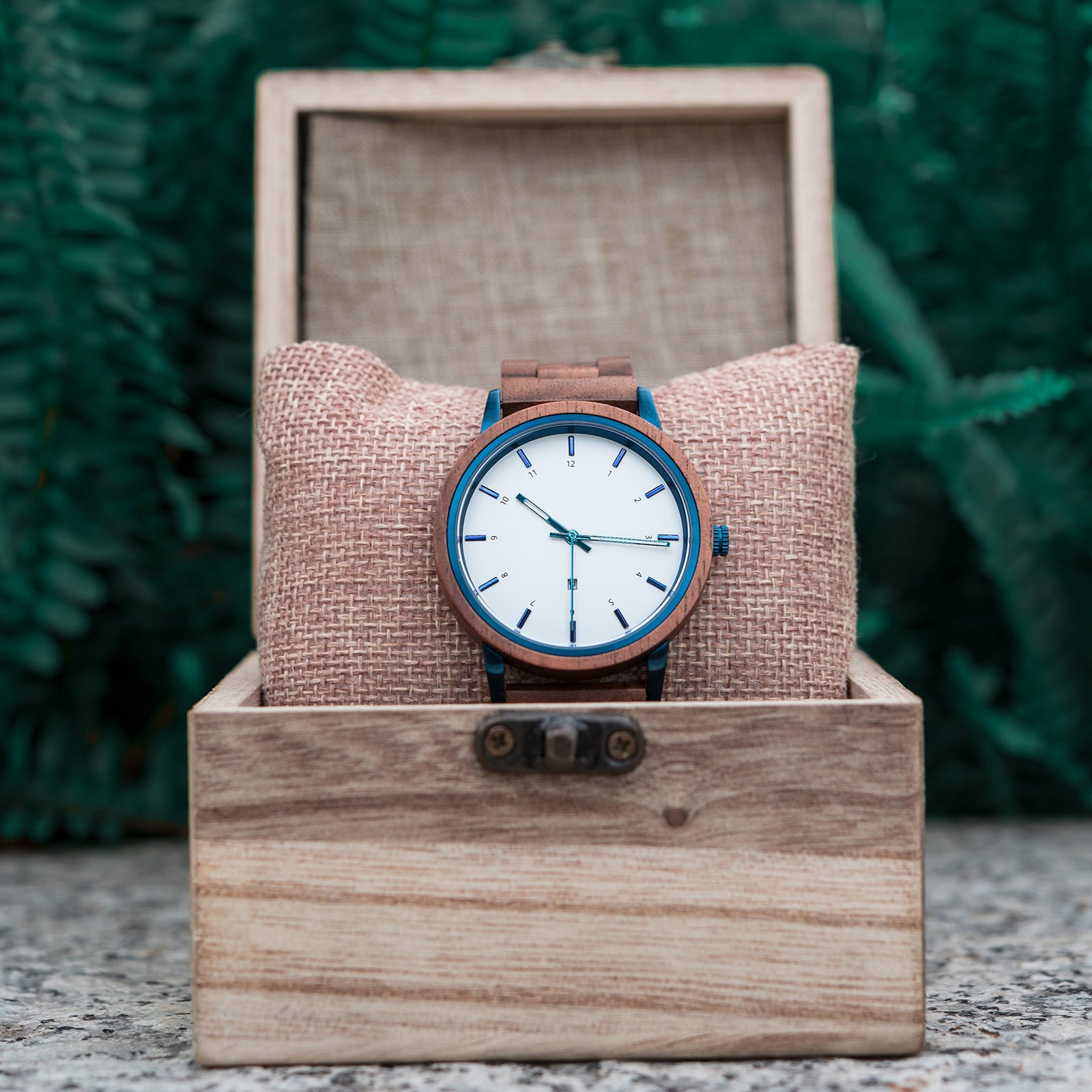 A stylish wooden wristwatch with a white dial and blue accents, placed on a burlap cushion inside a wooden box, set against a backdrop of green ferns.