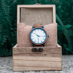 A stylish wooden wristwatch with a white dial and blue accents, placed on a burlap cushion inside a wooden box, set against a backdrop of green ferns.