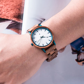 A wooden wristwatch with a white dial and teal accents worn on a person's wrist, featuring a natural wood band and a stylish design.
