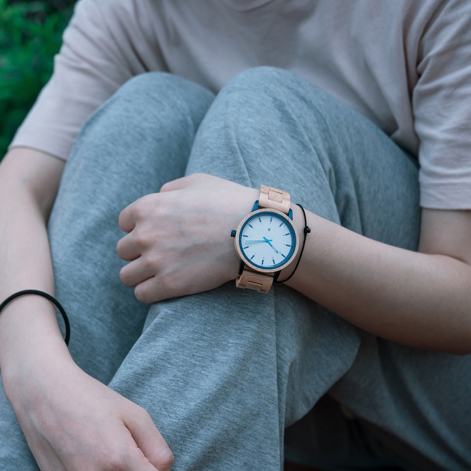 A person wearing a stylish wooden wristwatch with a white dial and blue accents, paired with casual gray clothing, in an outdoor setting with greenery.