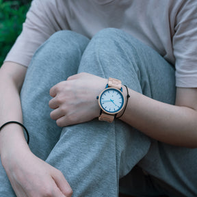 A person wearing a stylish wooden wristwatch with a white dial and blue accents, paired with casual gray clothing, in an outdoor setting with greenery.