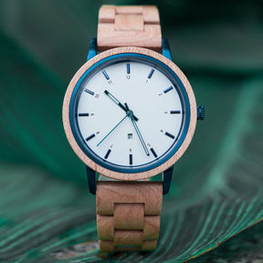 A close-up of a stylish wooden wristwatch with a white dial, blue accents, and a natural wood band, placed on green fern leaves.